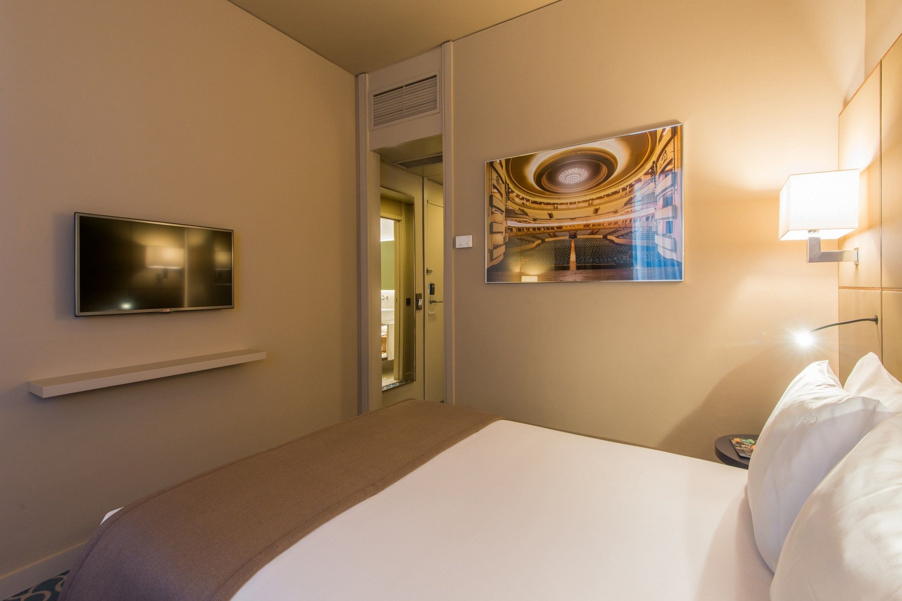 Ein Hotelzimmer mit Bett, Wandfernseher, Gemälde und Nachttischlampe.