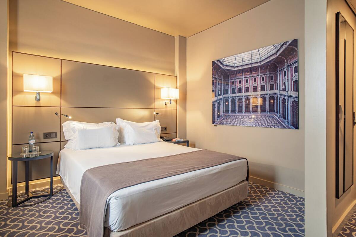 A hotel room with a large bed, two bedside lamps, a nightstand, a water bottle, glasses, and a framed picture of a historic building.