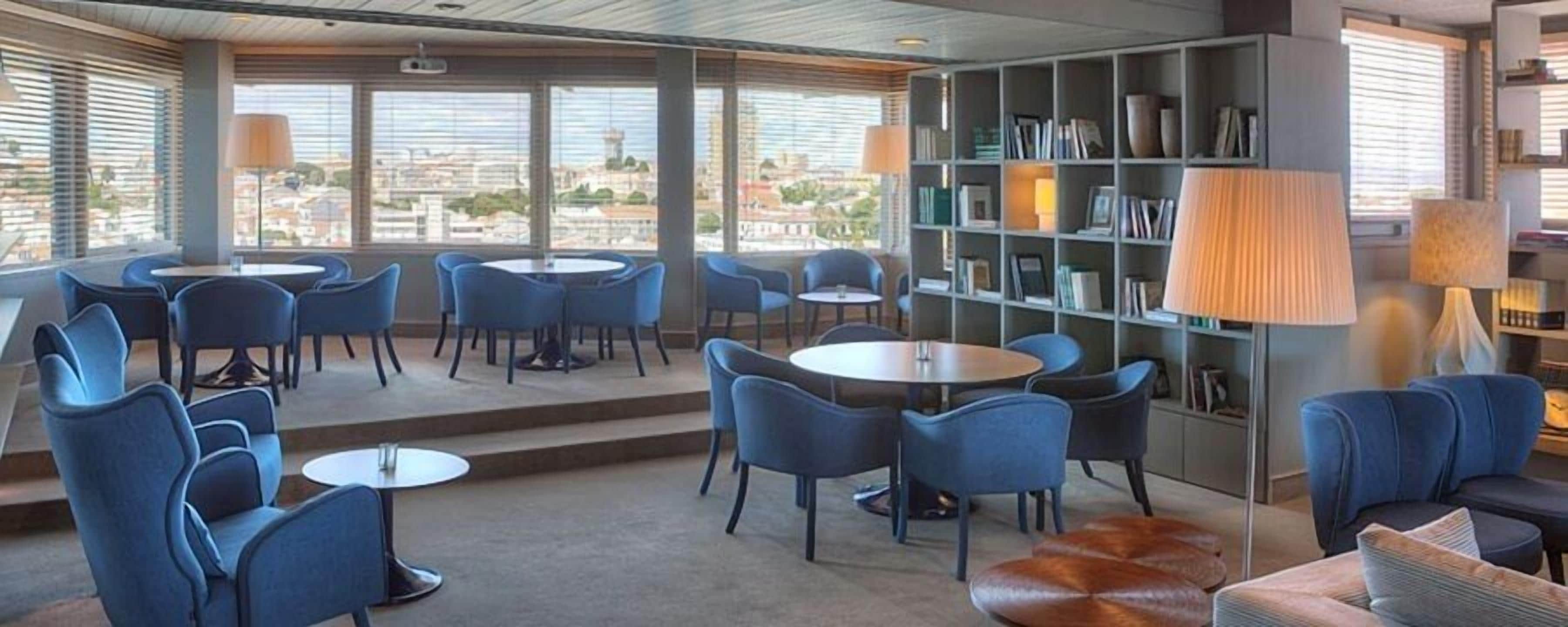 A modern lounge area with blue chairs and tables, a bookshelf, and large windows offering a city view.