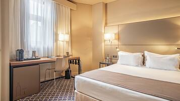 Habitación de hotel con cama, escritorio, silla y una mesita de noche con lámpara.