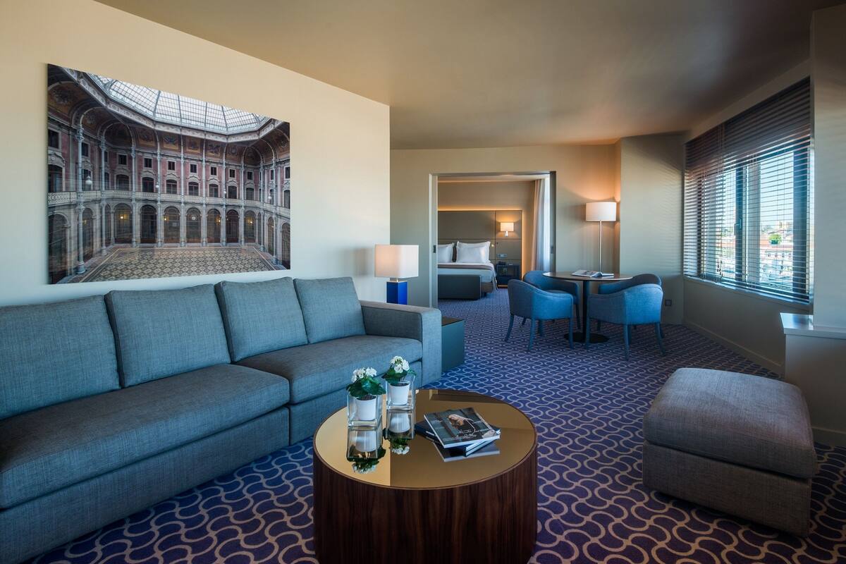 A hotel room with a grey sofa, a round coffee table with decorative items, a patterned carpet, a painting of a historical building, and a view through a window with blinds.