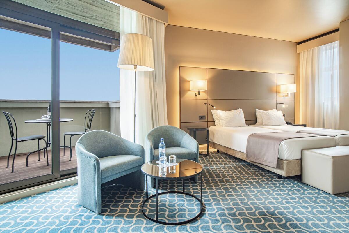 A hotel room with a bed, two armchairs, a small table with a bottle and glasses, and a view of the outdoors.
