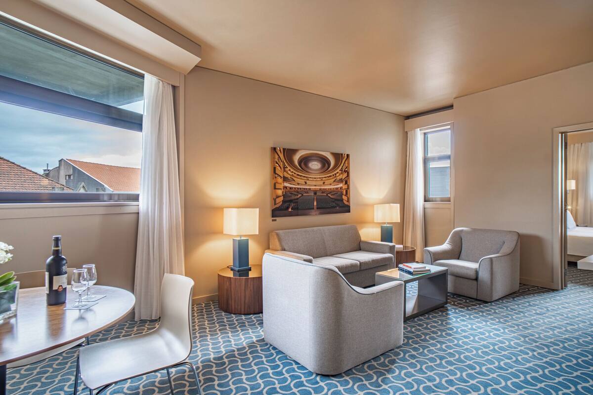 A hotel room with a round table, two armchairs, a sofa, a coffee table, and a large window with curtains.