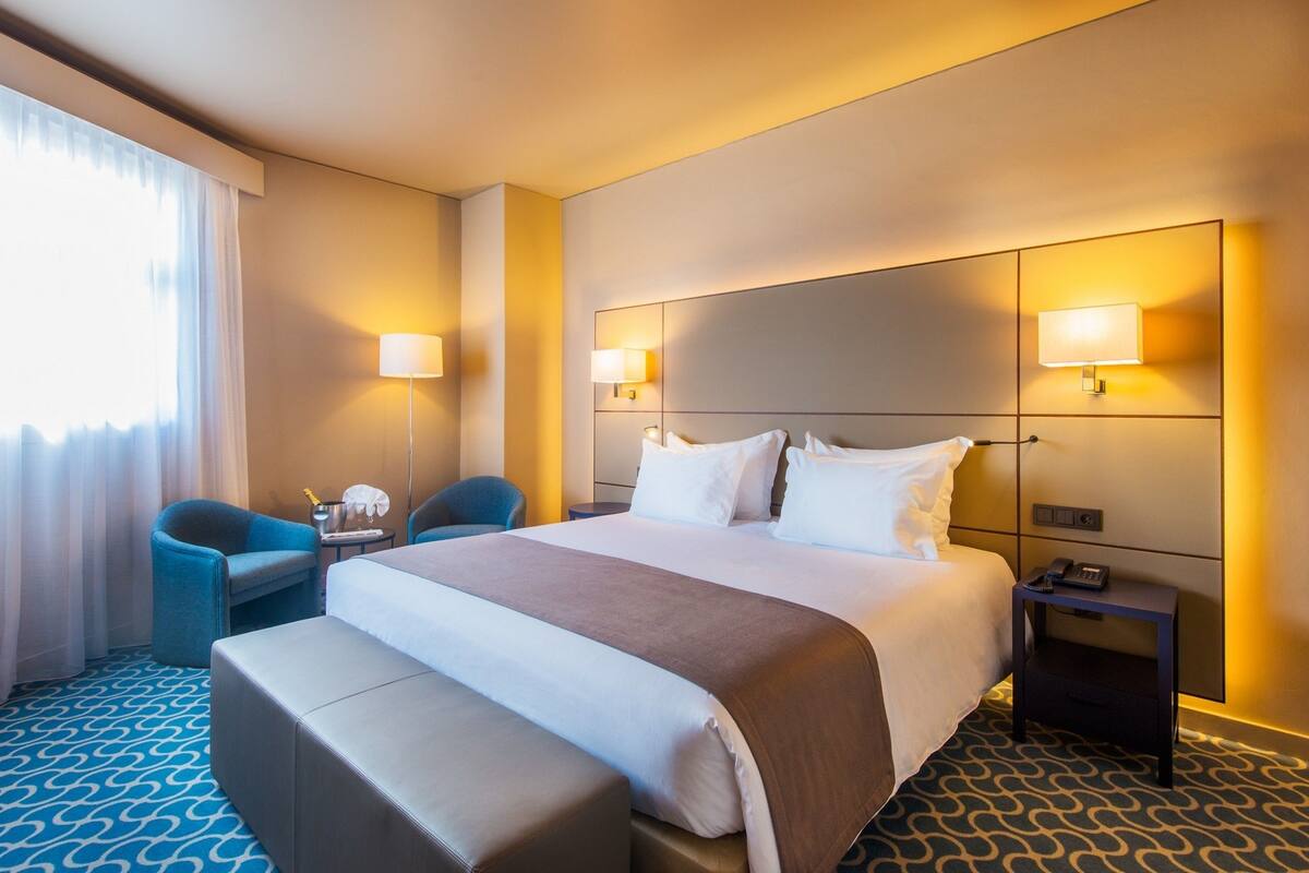 A hotel room with a large bed, two bedside lamps, a small table with a phone, a chair, and a sofa.
