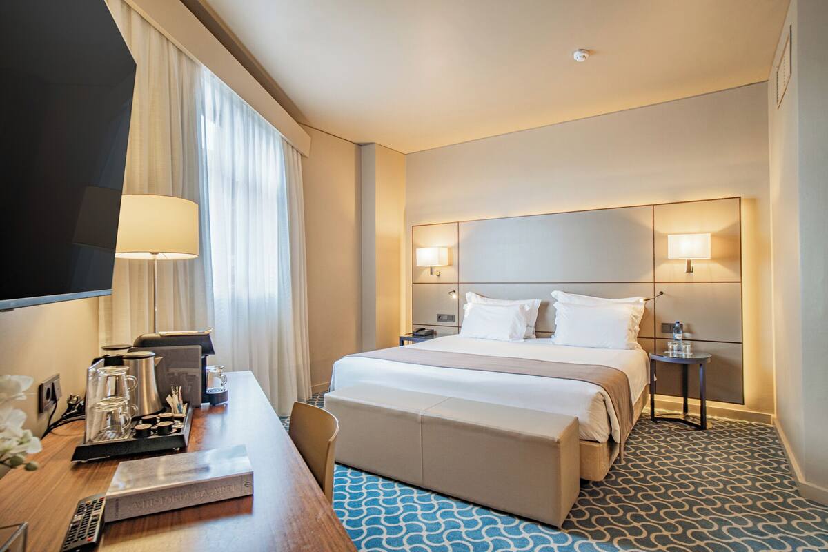 A hotel room with a large bed, bedside lamps, a desk with a coffee maker, and a TV.