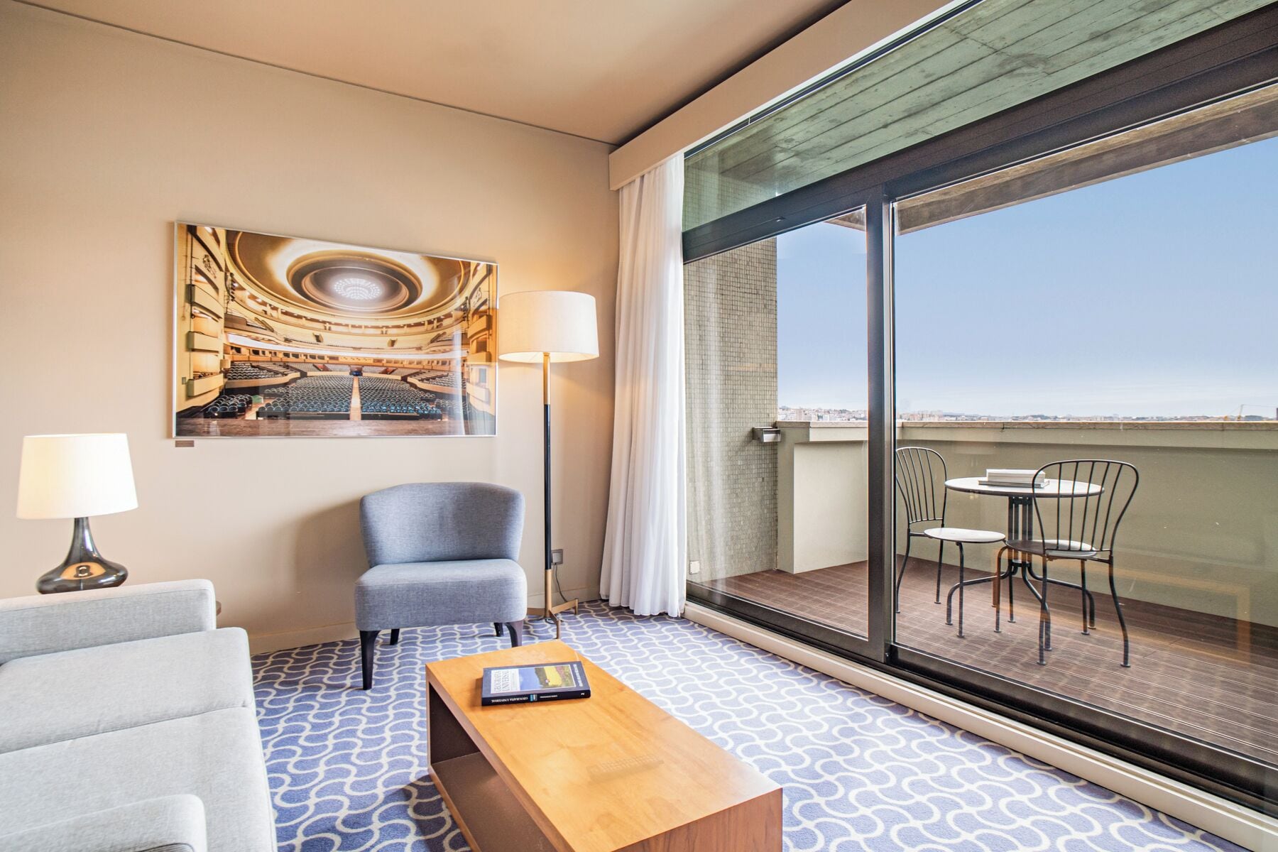 Habitación de hotel con un ventanal grande, un sofá, una silla, una mesa y un cuadro de un auditorio.