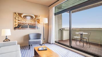Habitación de hotel con un ventanal grande, un sofá, una silla, una mesa y un cuadro de un auditorio.