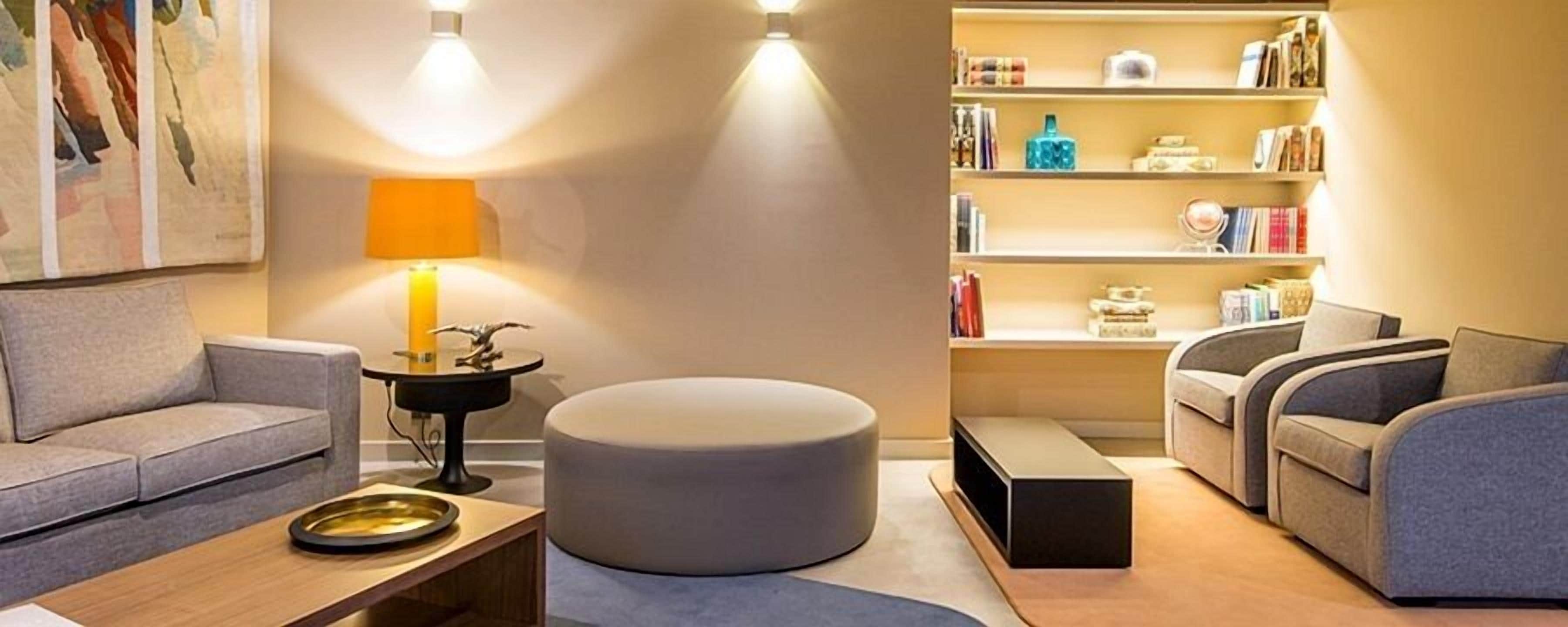 A modern living room with a grey sofa, a round ottoman, a wooden coffee table, and a bookshelf filled with books and decorative items.