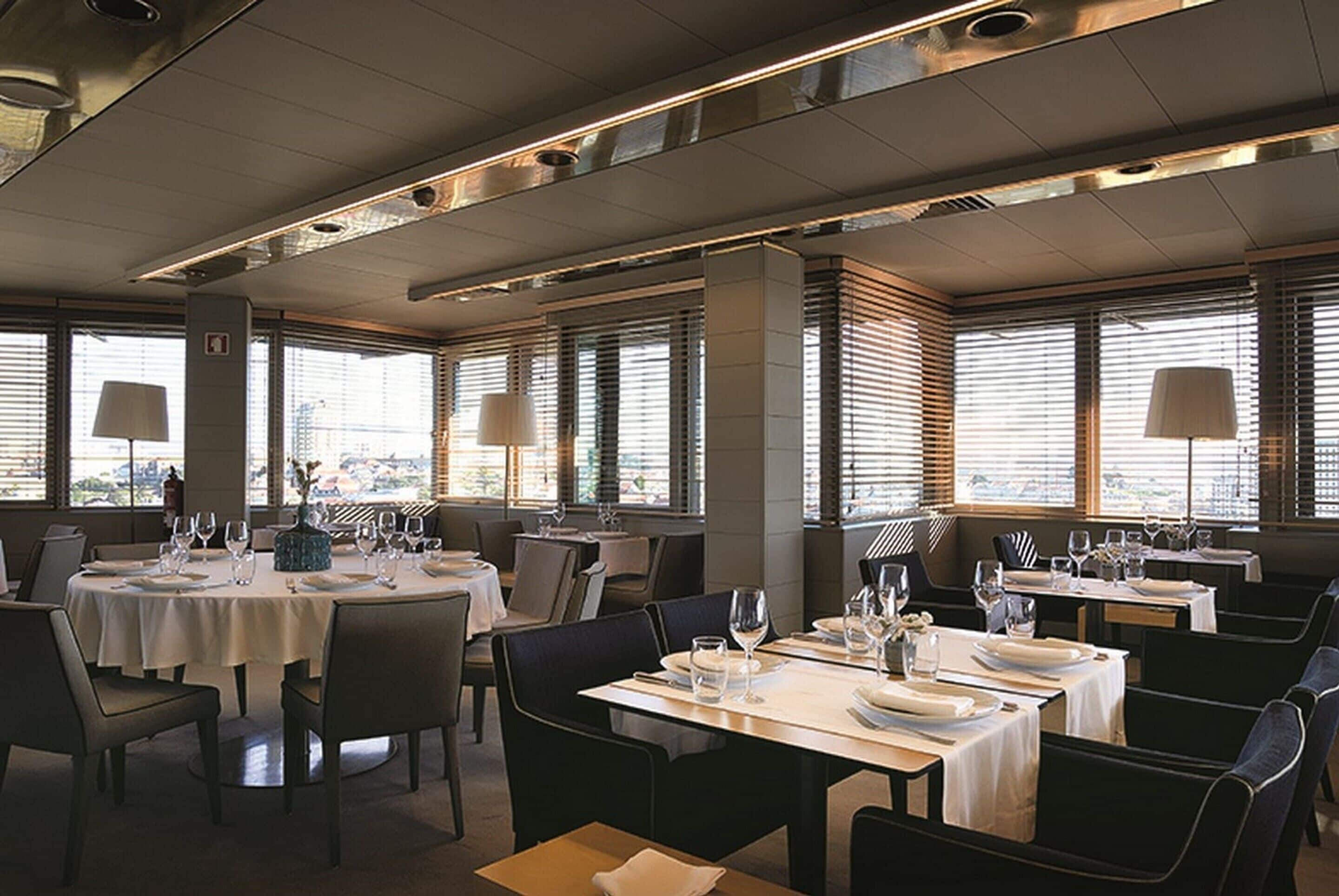 A restaurant with round tables set for dining, large windows with blinds, and modern lighting fixtures.