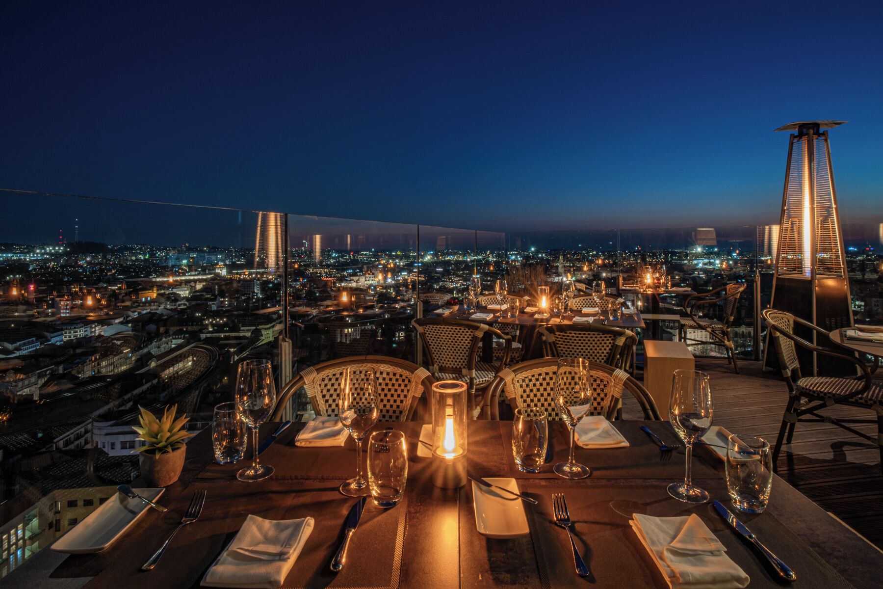 Une table à manger installée sur un toit, avec des chaises, une bougie allumée et une vue sur la ville au crépuscule.