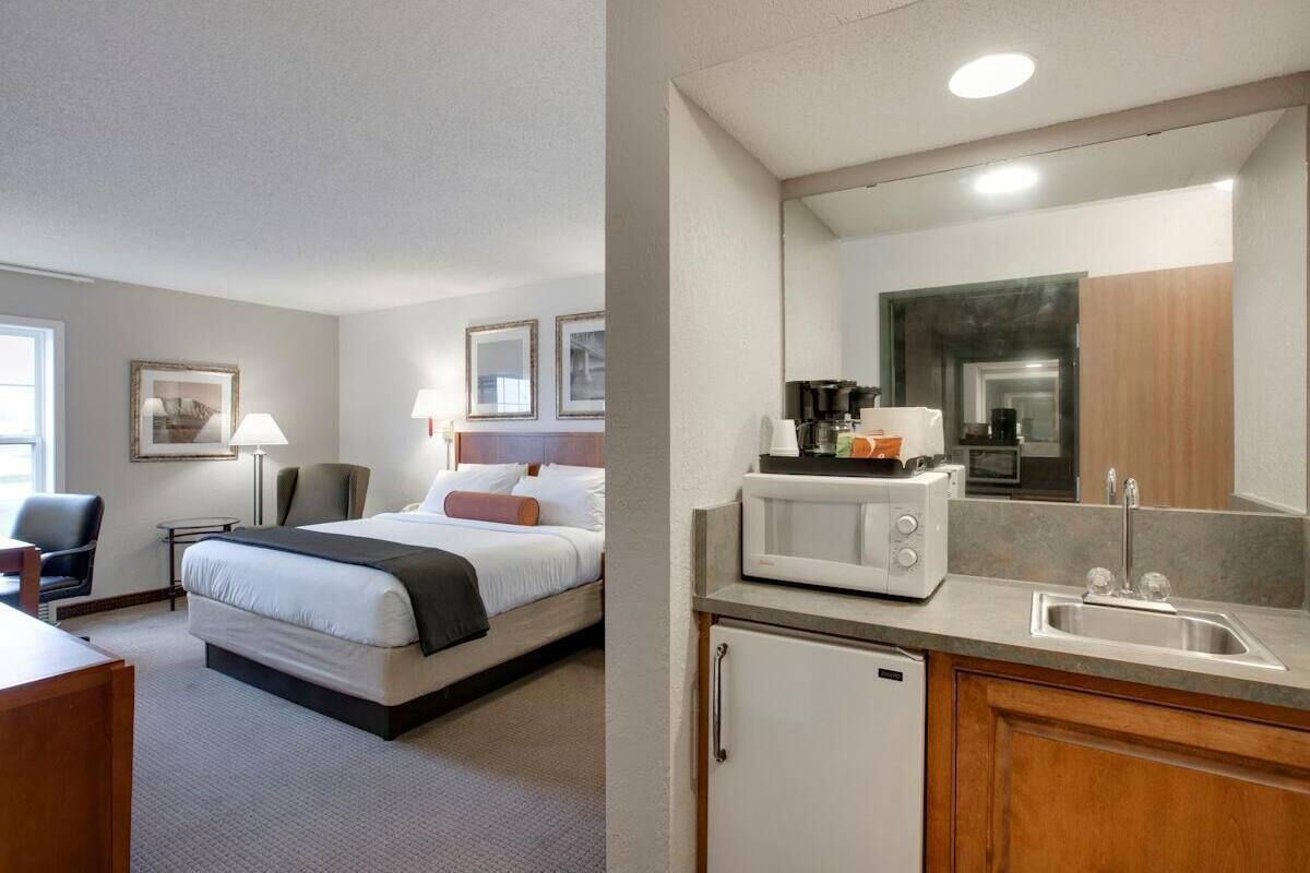 A hotel room with a bed, bedside table, lamp, chair, and a kitchenette with a microwave, coffee maker, and sink.