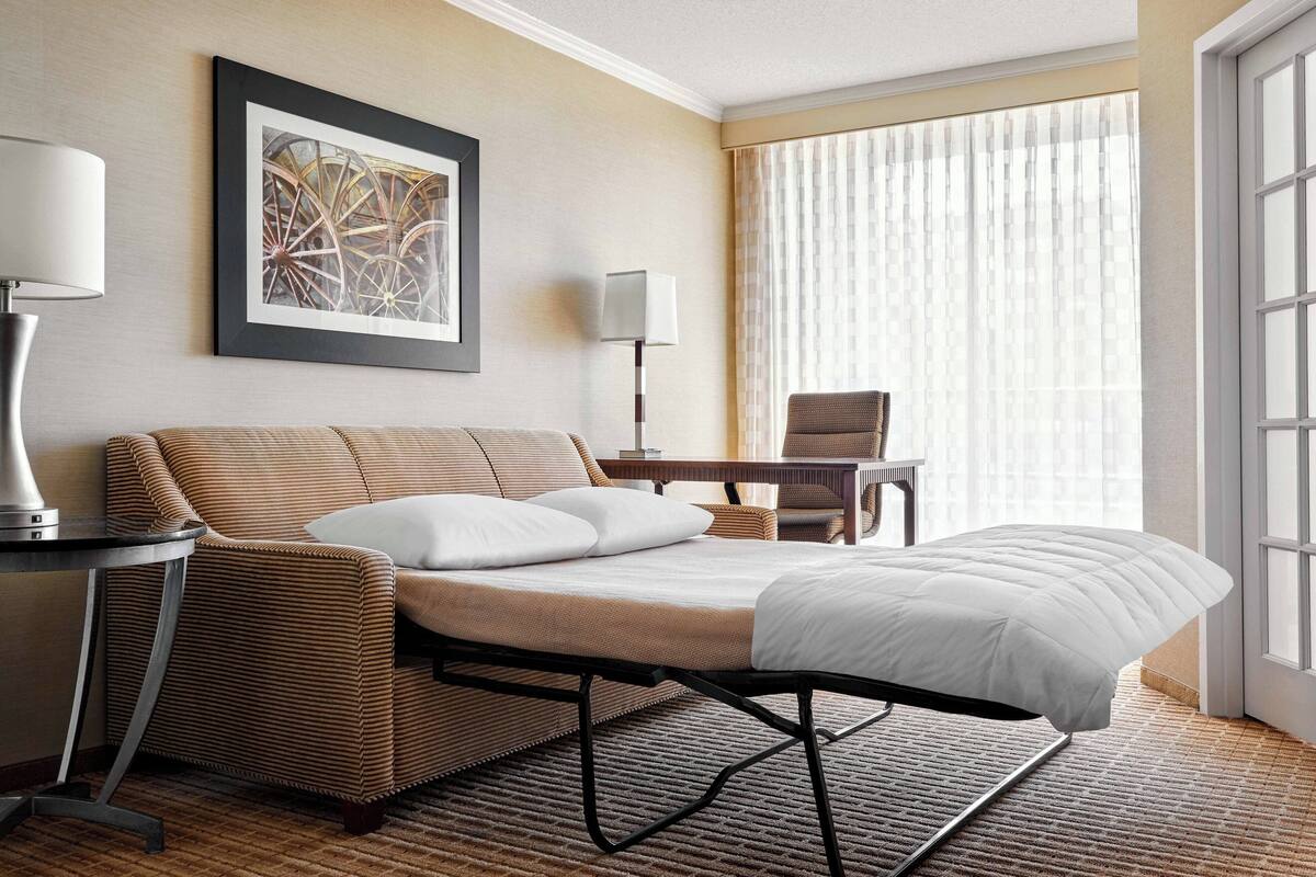 Quarto de hotel com cama, cadeira, escrivaninha e uma janela grande com cortinas.