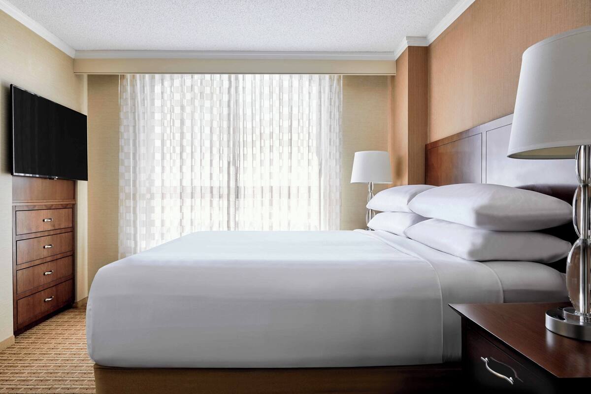Quarto de hotel com uma cama grande, televisão de tela plana, cômoda de madeira e janela com cortinas translúcidas.