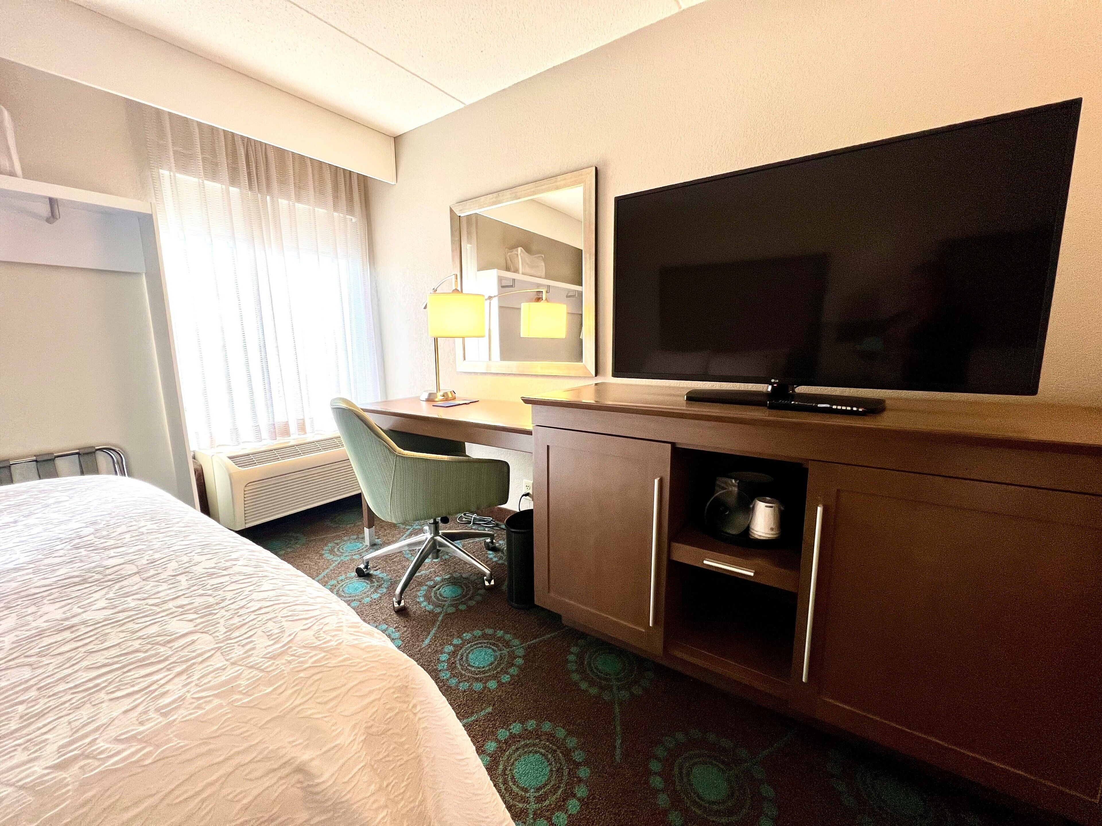 A hotel room with a bed, a desk with a chair, a television, a mirror, and a window with curtains.