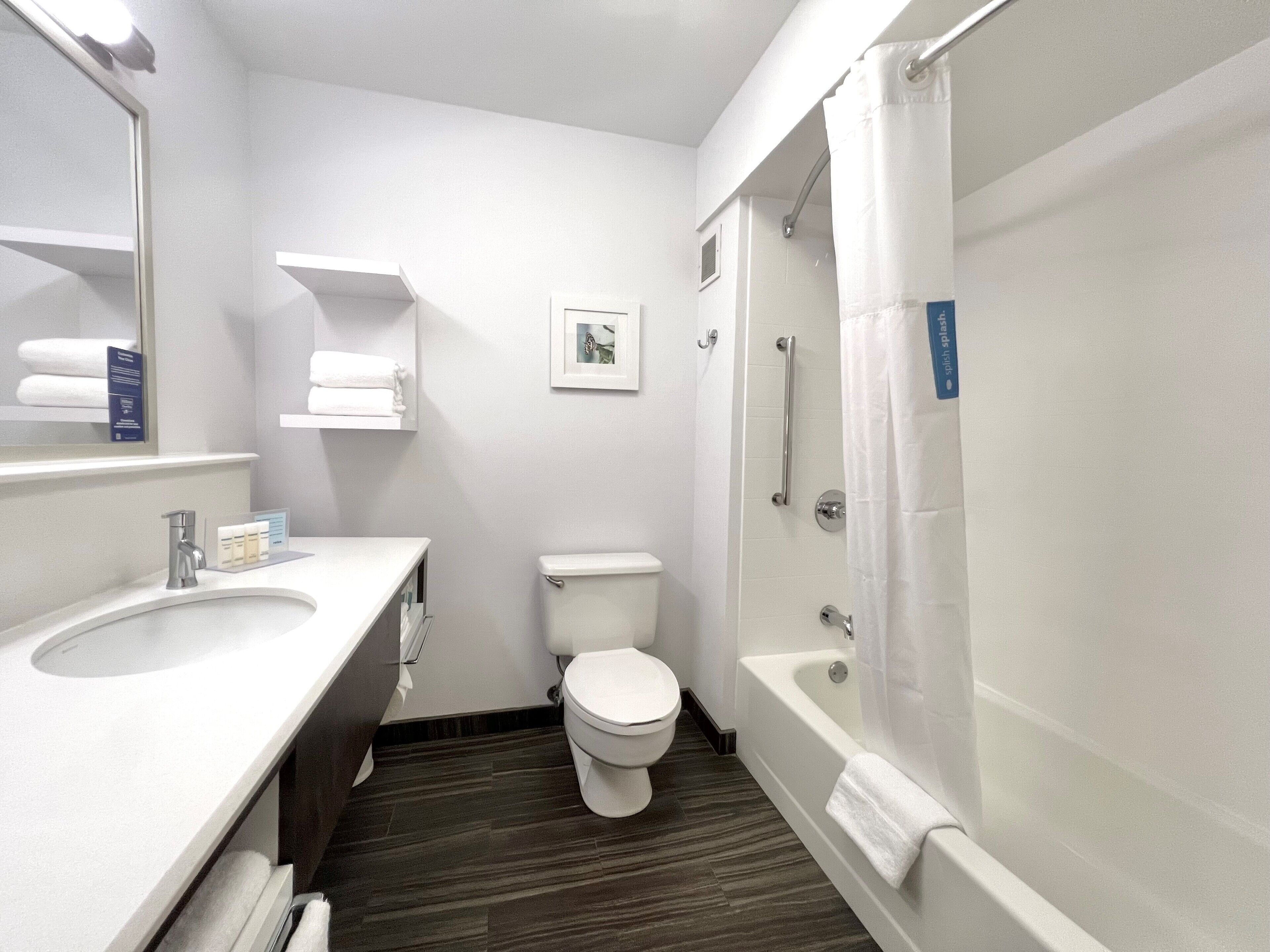 A modern bathroom with a white sink, toilet, and bathtub. There are shelves with towels and a framed picture on the wall.
