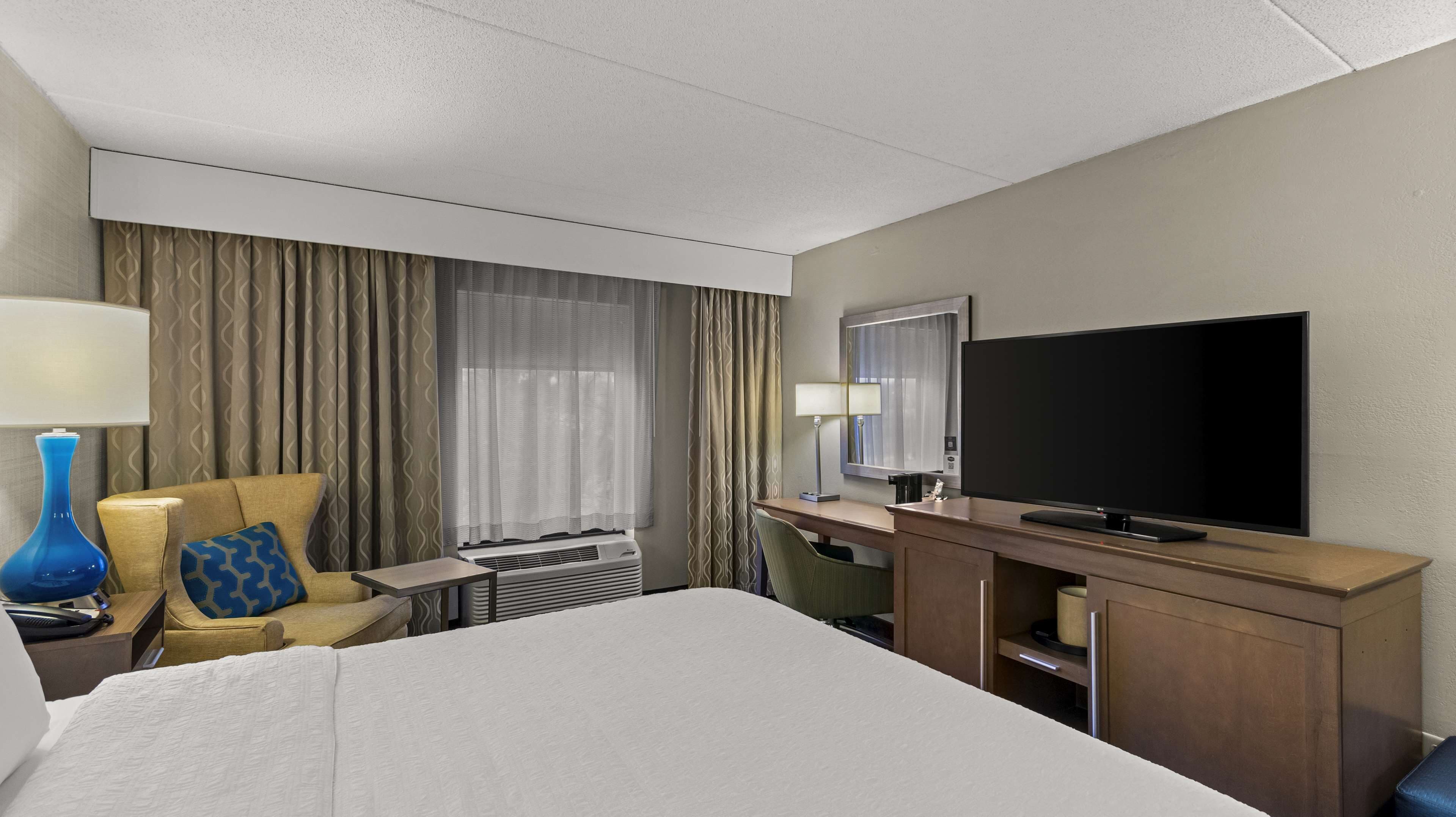 Habitación de hotel con cama, escritorio, televisor de pantalla plana, silla, lámpara y ventana con cortinas.