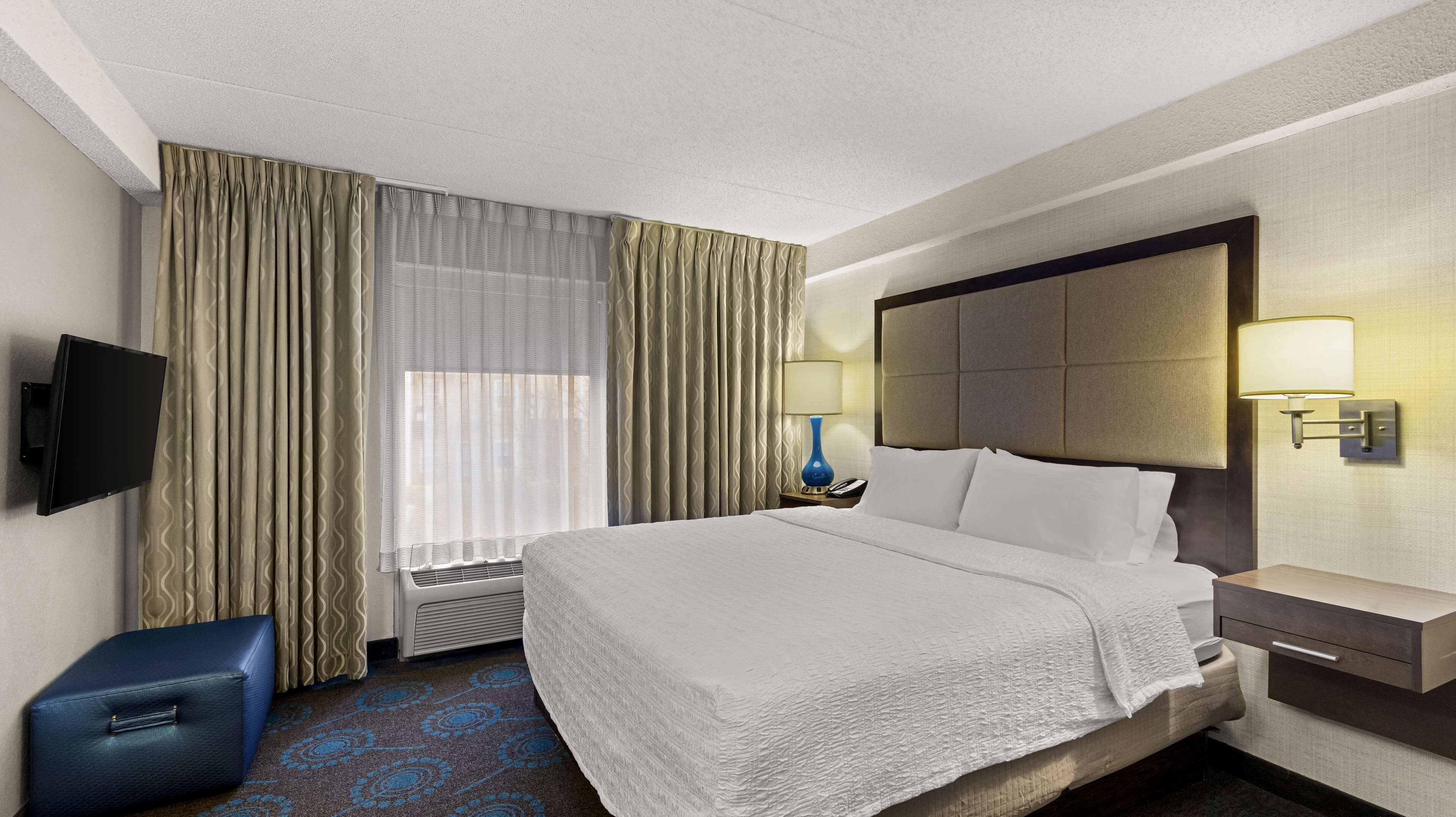 Habitación de hotel con una cama grande, un televisor de pantalla plana, una lámpara de noche, una mesita de noche y una ventana con cortinas.