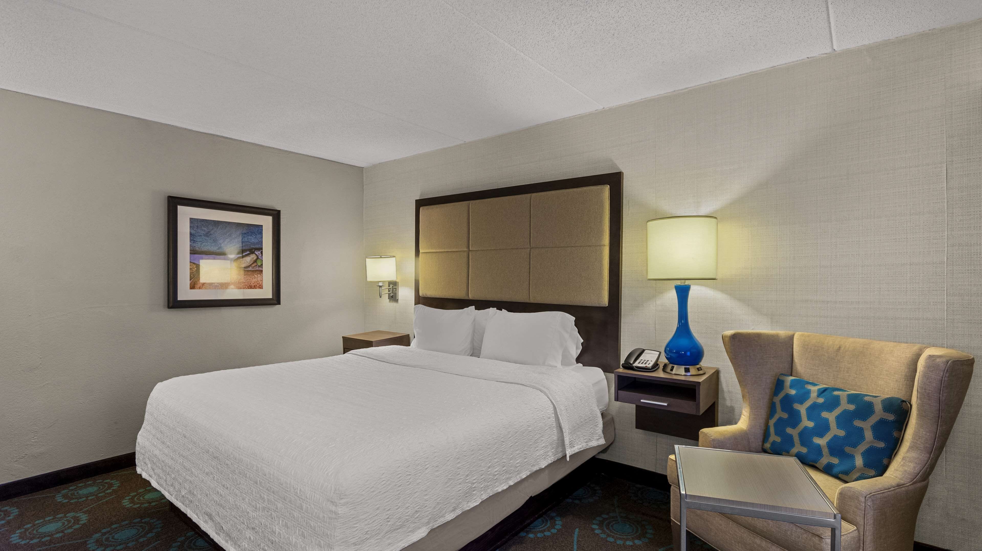 A hotel room with a large bed, a chair, a nightstand with a lamp, and a framed picture on the wall.