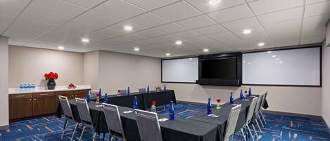 Una sala de conferencias con una larga mesa, sillas, una pantalla de proyector y una pizarra.