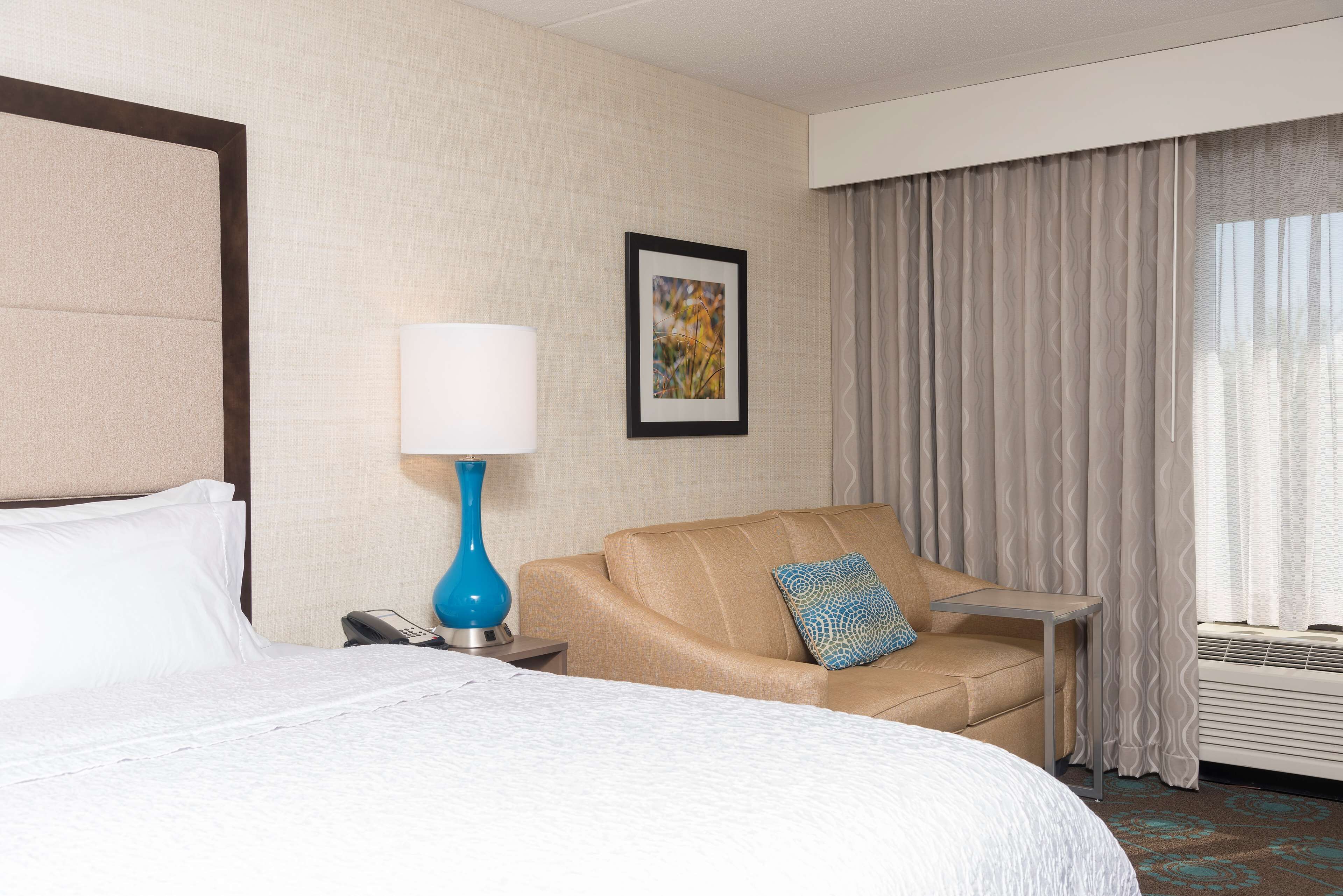 A hotel room with a bed, a sofa, a small table, a lamp, a phone, a picture on the wall, and a window with curtains.