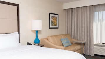 A hotel room with a bed, a sofa, a small table, a lamp, a phone, a picture on the wall, and a window with curtains.