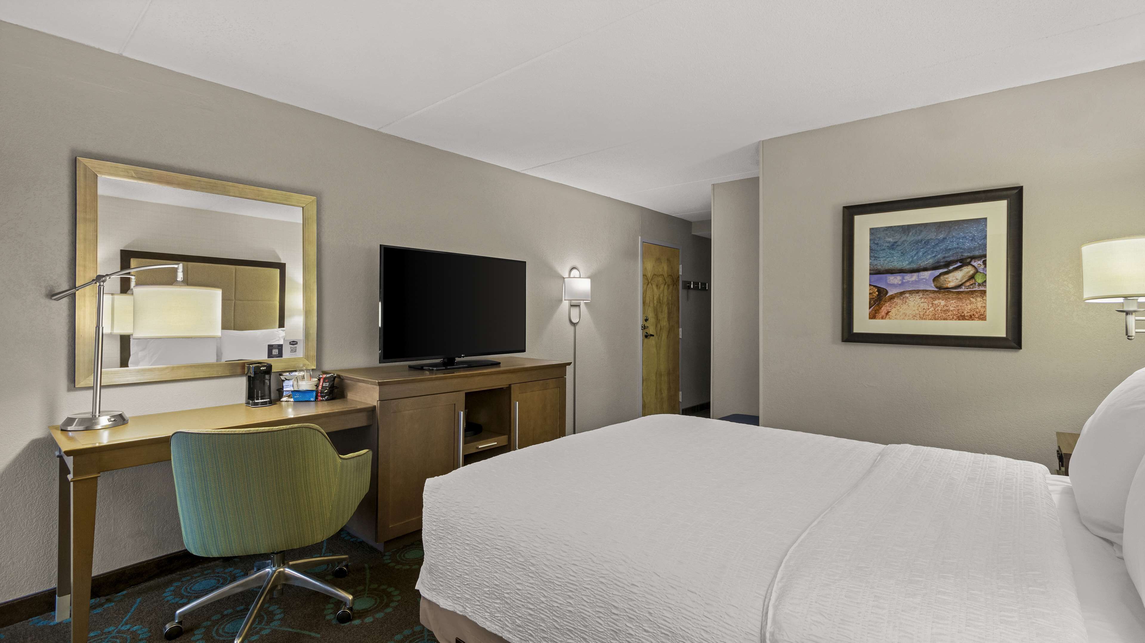 Une chambre d’hôtel avec un lit, un bureau avec un ordinateur, une chaise, une télévision, une lampe et un tableau au mur.