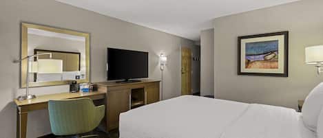 Una habitación de hotel con cama, escritorio con computadora, silla, televisor, lámpara y un cuadro en la pared.