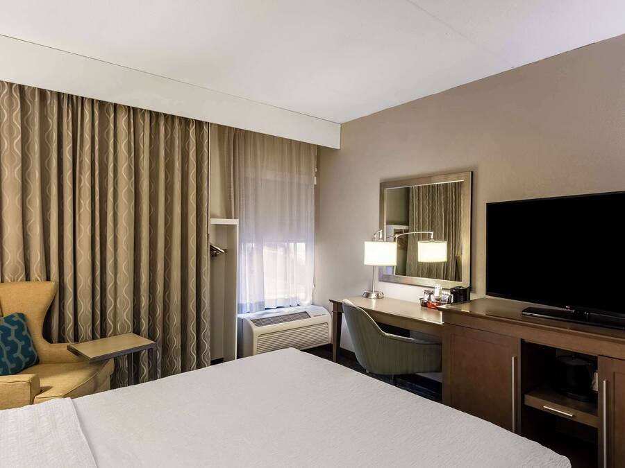 A hotel room with a bed, a desk with a TV, a chair, a desk lamp, and a large window with curtains.