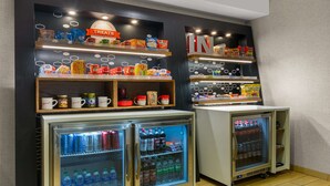 A snack and beverage area with shelves stocked with various snacks, a refrigerator with drinks, and a vending machine. - Hampton Inn Bloomington (Bloomington)