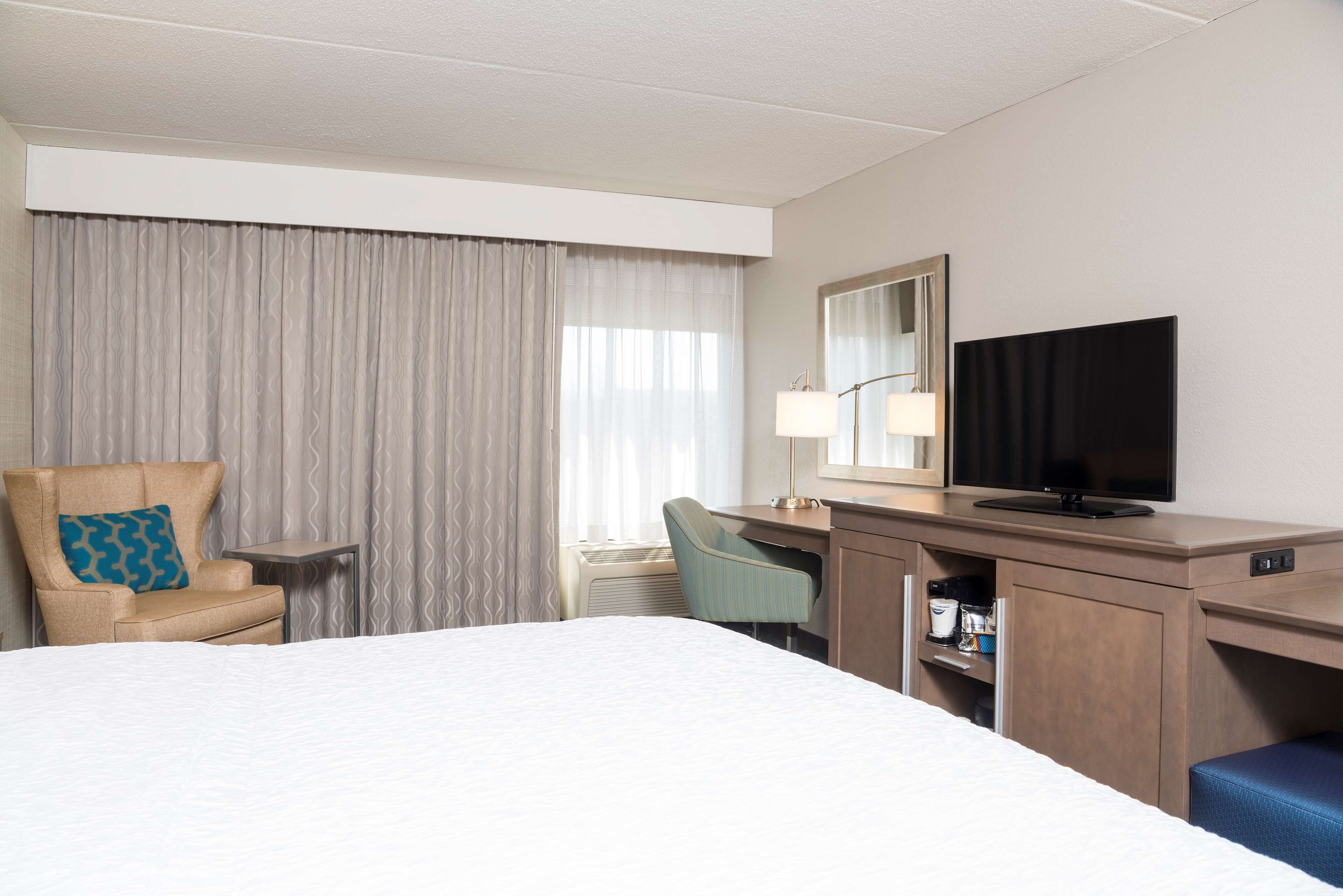 A hotel room with a bed, a desk with a television, a chair, and a desk lamp.