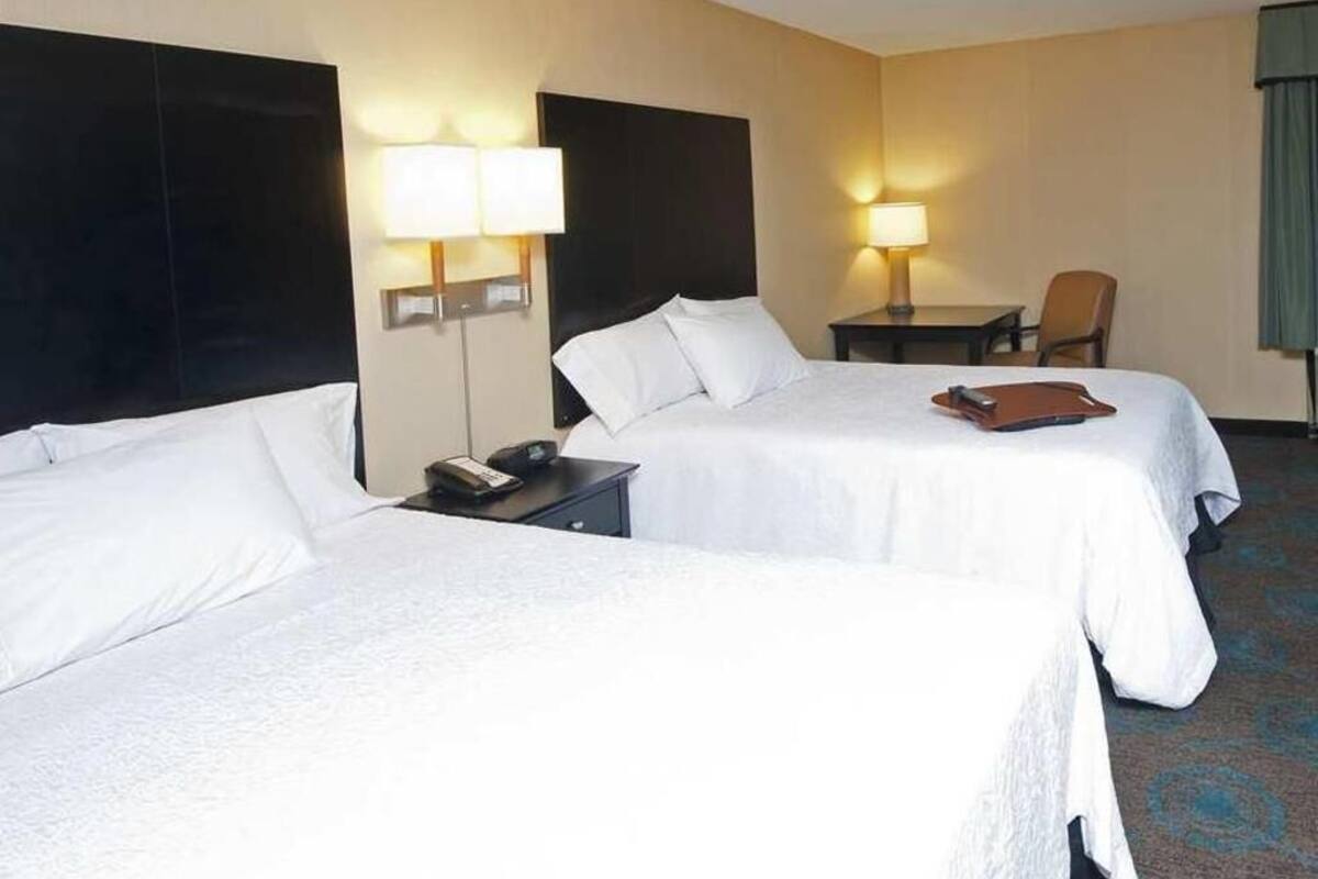 A hotel room with two beds, a desk, a chair, and a lamp.