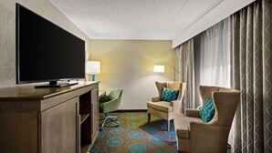A hotel room with a flat-screen TV, a desk with a lamp, two armchairs with patterned cushions, a patterned carpet, and a large window with curtains. - Hampton Inn Bloomington (Bloomington)