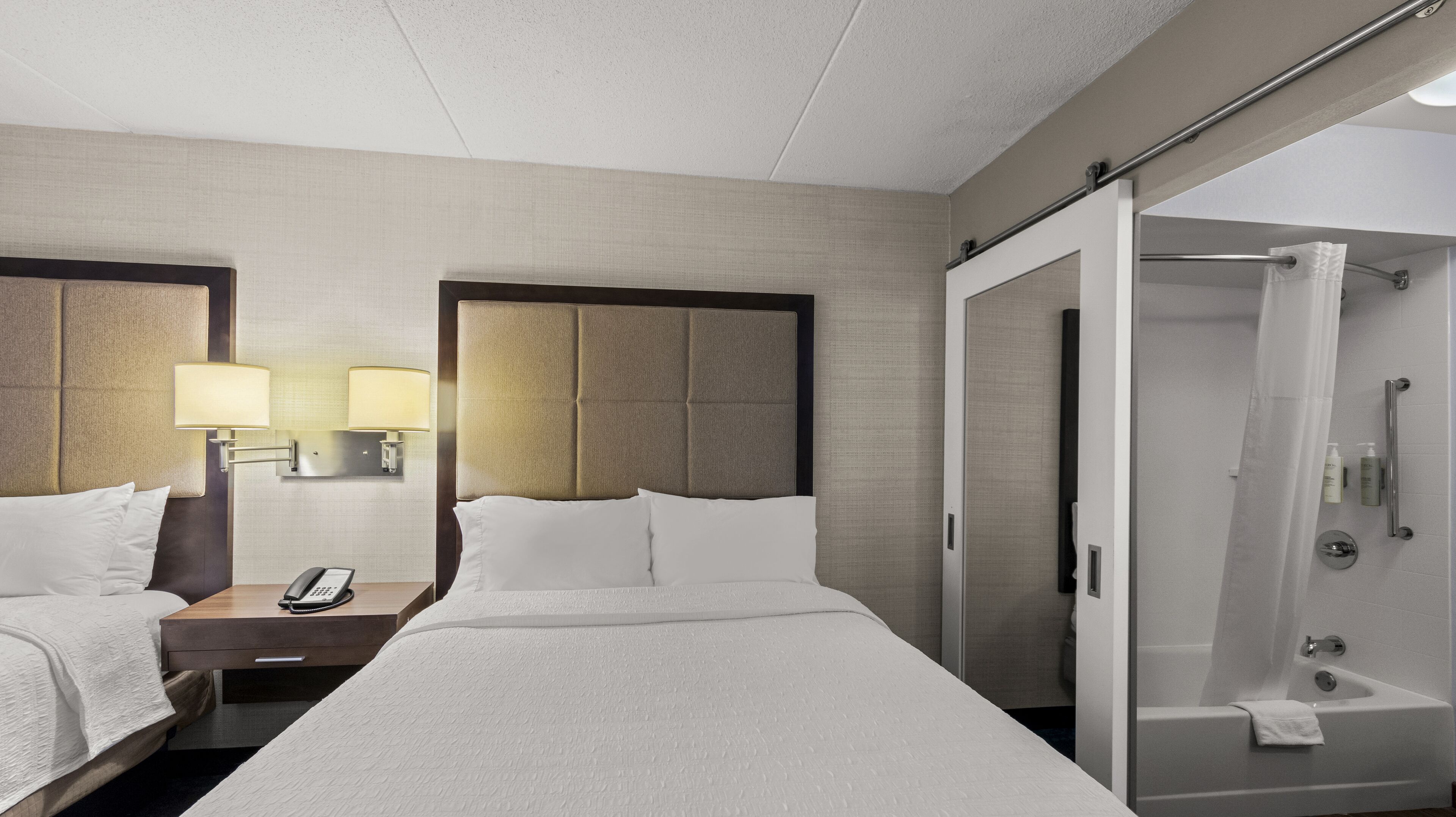 Habitación de hotel con una cama grande, dos mesitas de noche con lámparas, zona de ducha y baño.