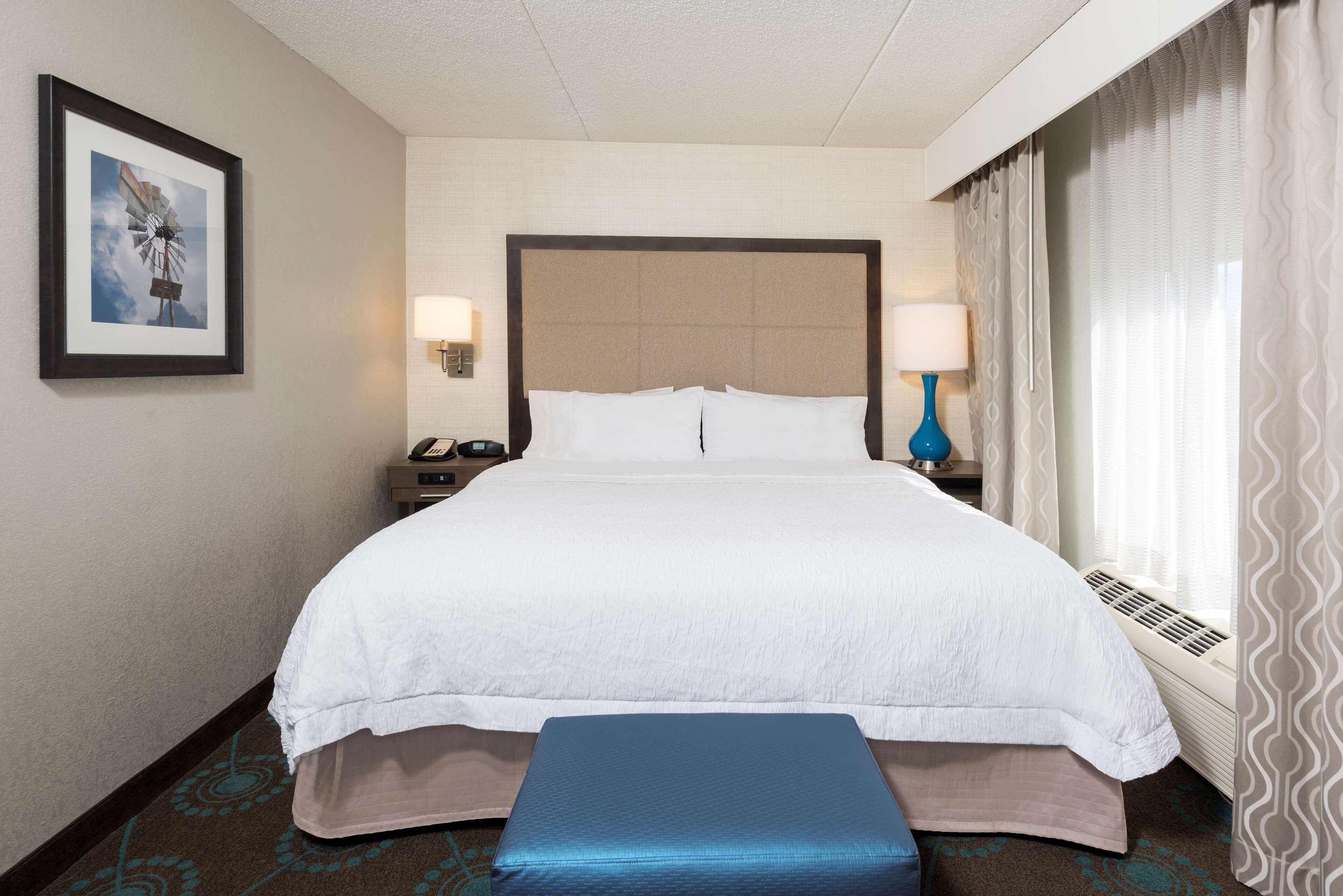A hotel room with a large bed, a bedside table with a lamp, a framed picture on the wall, and a window with curtains.