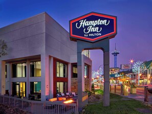 A Hampton Inn by Hilton hotel with a prominent sign, outdoor seating area, and a view of the cityscape at dusk.