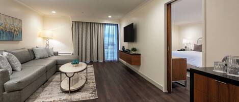 A modern hotel room with a grey sofa, a round coffee table, a wooden dresser, a bed with a white duvet, and a TV mounted on the wall.