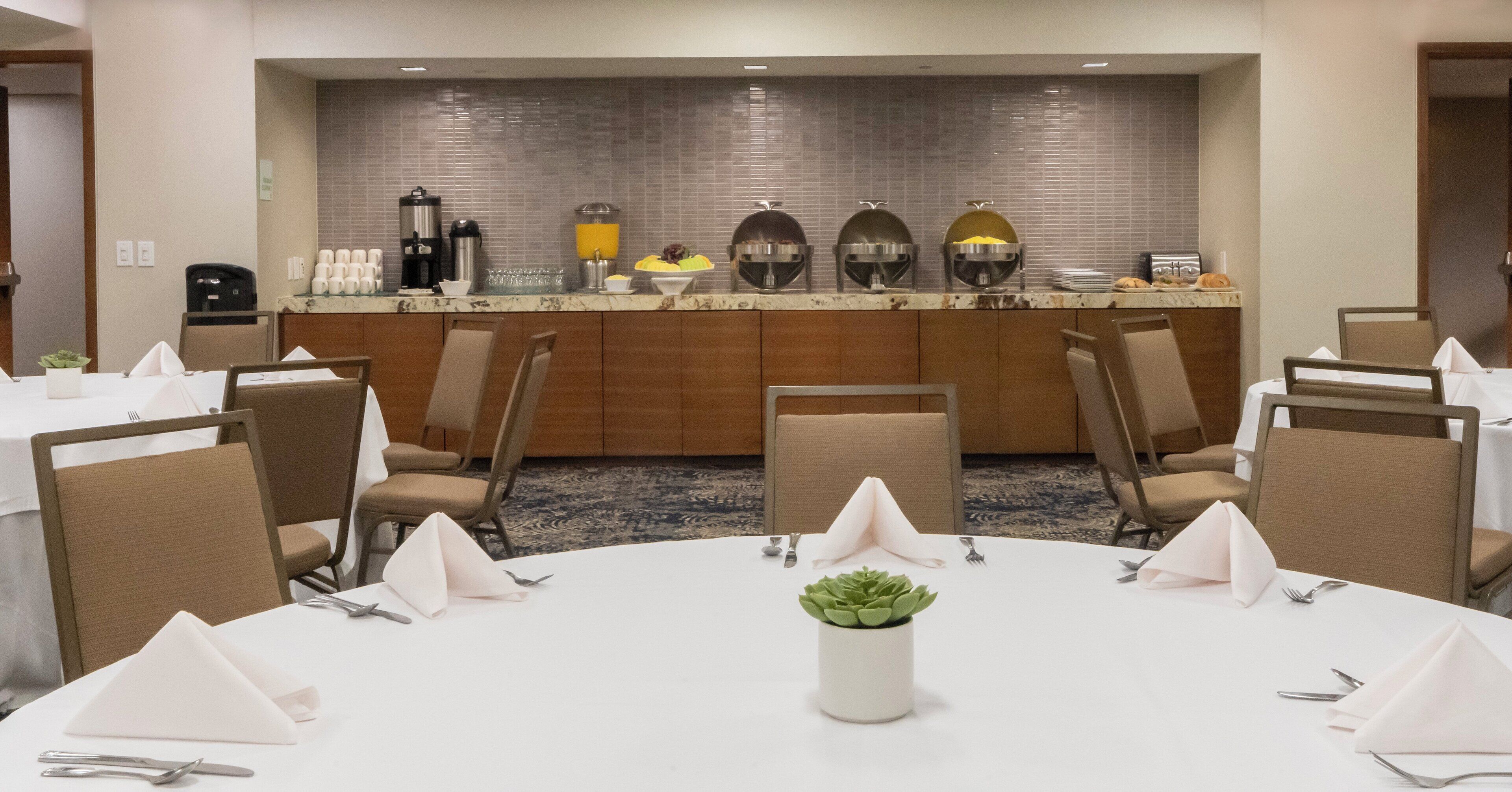 A conference room with round tables, chairs, and a buffet area with food and drink dispensers.