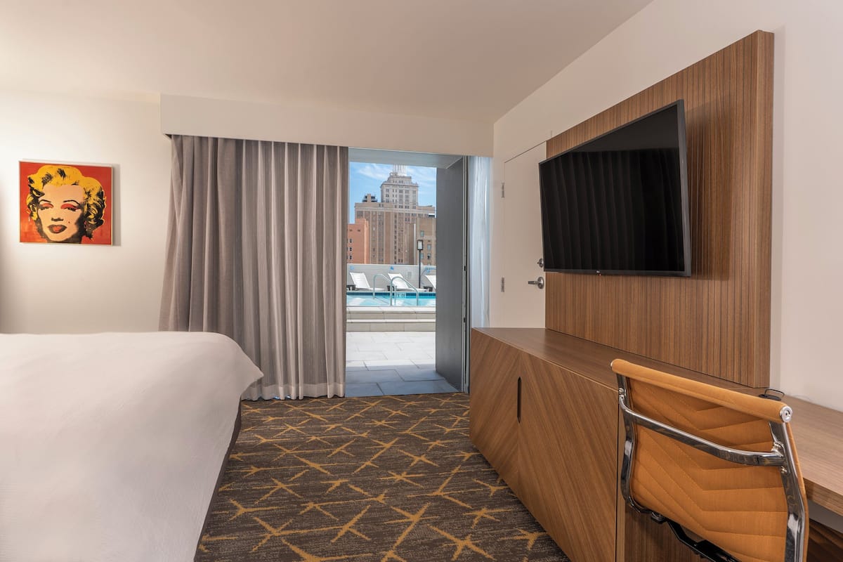 A hotel room with a bed, a desk with a TV, a chair, a large window with curtains, and a city view.