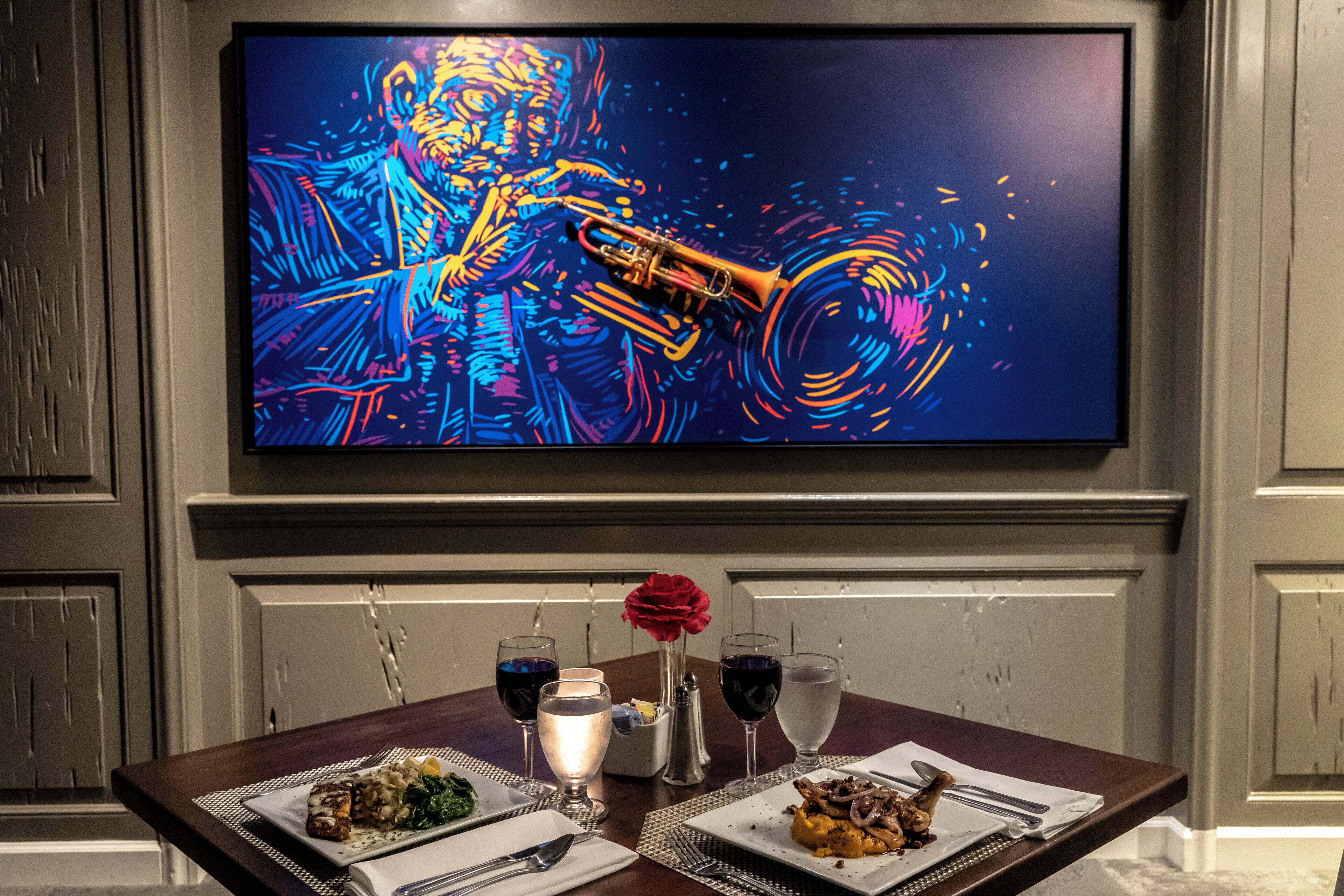 Une table à manger est dressée avec des assiettes remplies de nourriture, des verres et des couverts. Au-dessus, un téléviseur diffuse une image colorée.