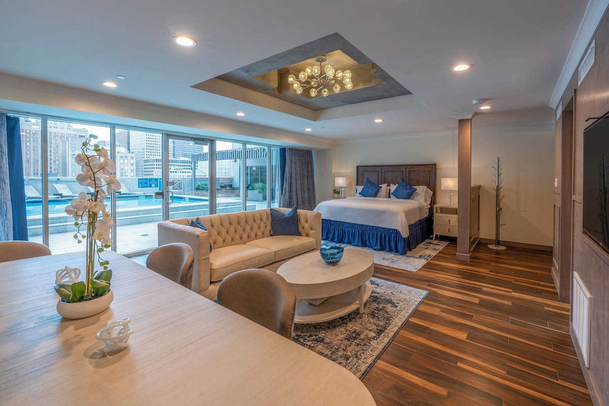 A modern hotel room with a large bed, a sofa, a dining table, and a view of the city through large windows.