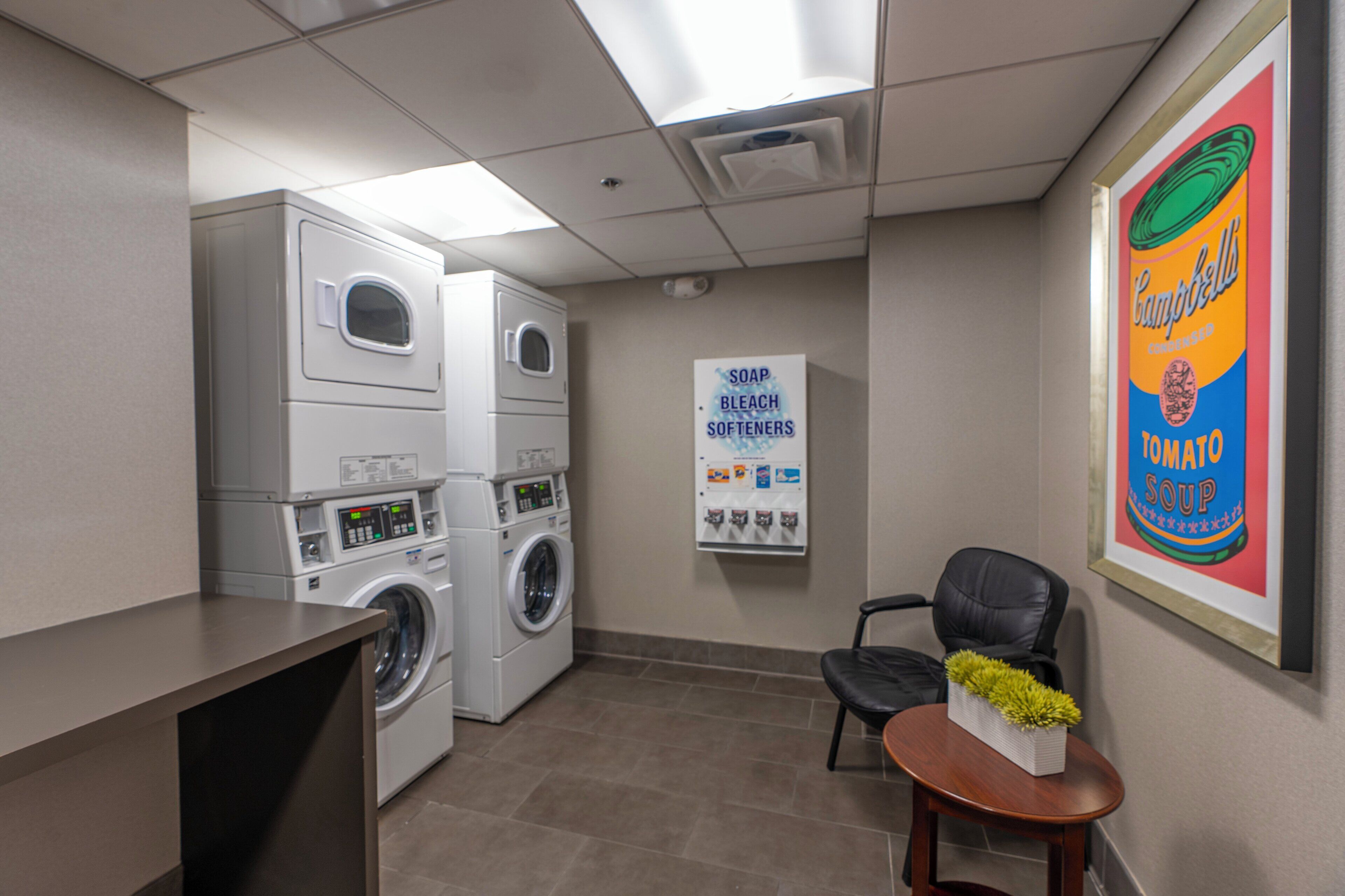 A laundry room with stacked washers and dryers, a sign listing available detergents, and a framed Campbell's soup advertisement.