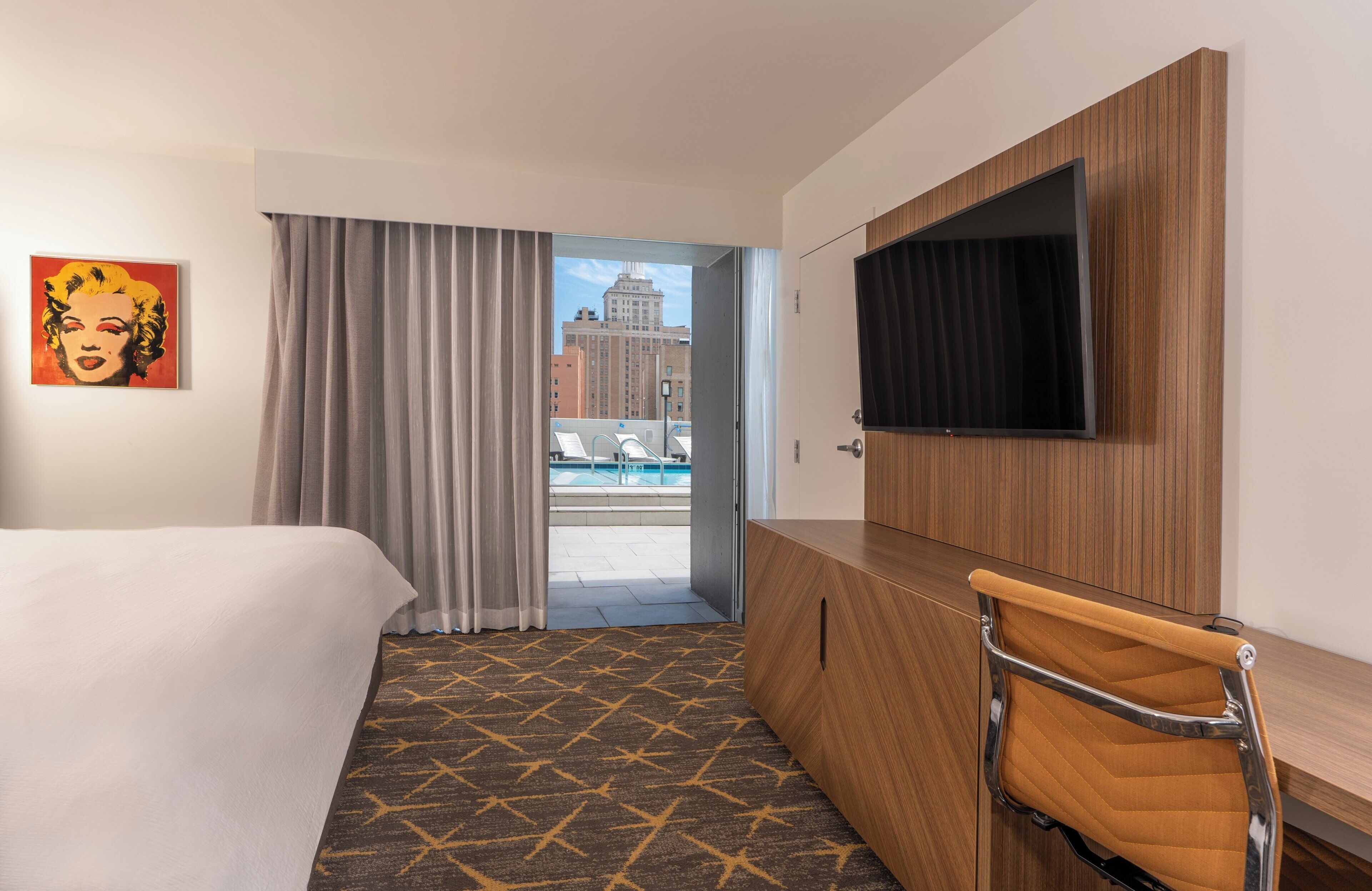 Habitación de hotel con cama, escritorio con televisor, una silla, ventana grande con cortinas y vista a la ciudad.