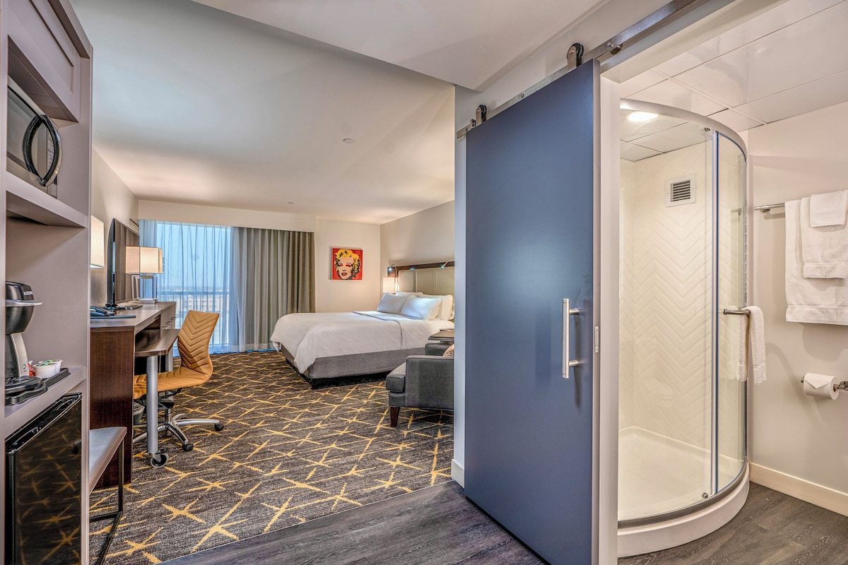 Habitación de hotel con cama, escritorio, silla y zona de ducha.