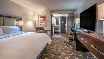 A hotel room with a large bed, a desk with a flat-screen TV, a chair, a bathroom with a mirror, and a patterned carpet.