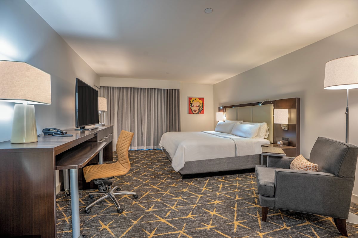 A hotel room with a bed, desk, chair, and a large window with curtains.