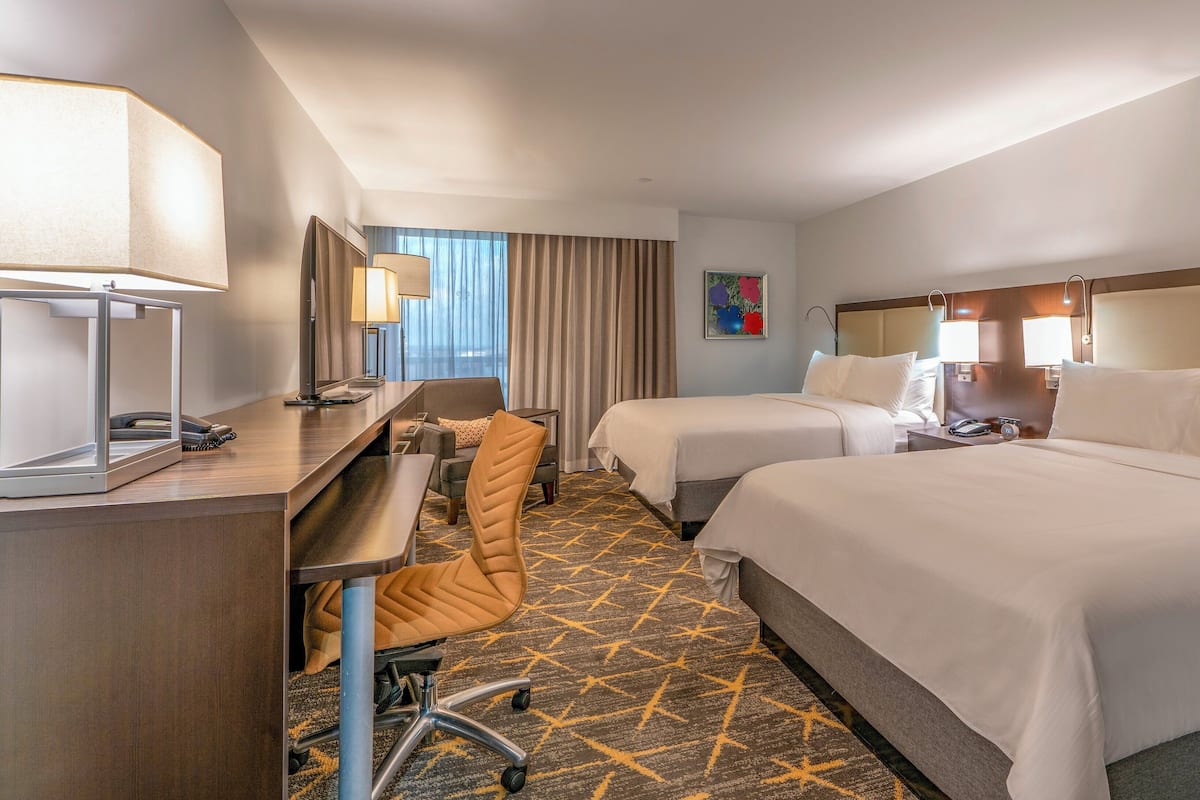 A hotel room with two beds, a desk, a chair, and a large window with curtains.