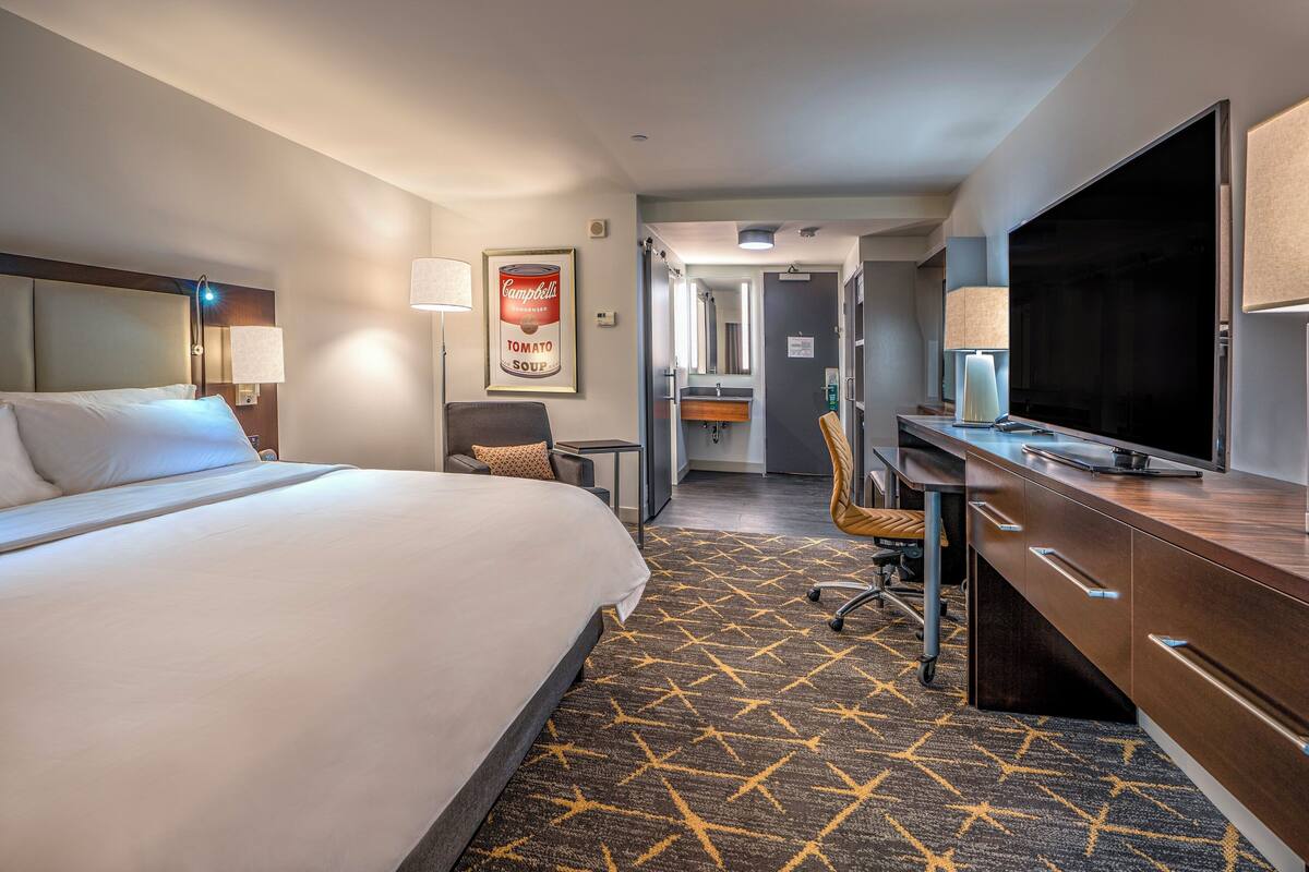 A hotel room with a large bed, a desk with a flat-screen TV, a chair, a bathroom with a mirror, and a patterned carpet.
