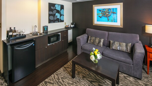 A modern living room with a gray sofa, a coffee table with flowers, a kitchen area with a dishwasher, and artwork on the walls. - Cambridge Suites Hotel (Halifax)