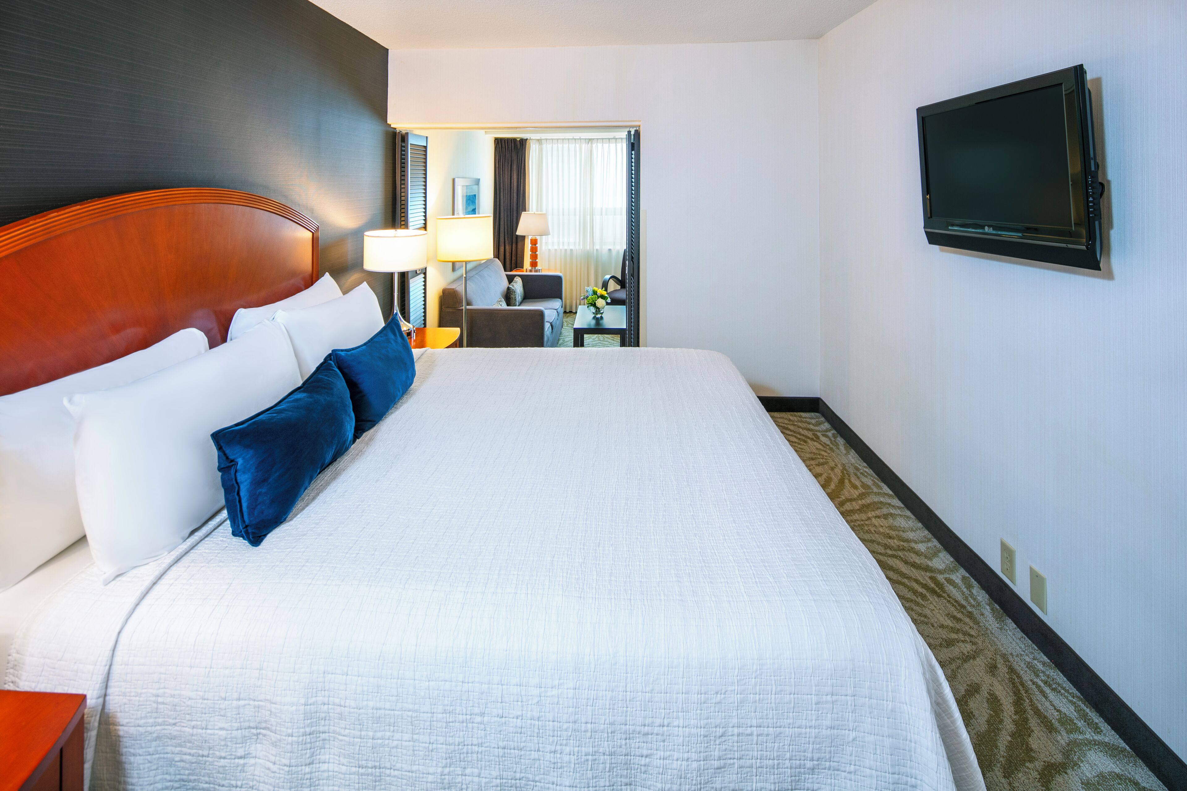 Une chambre d’hôtel avec un grand lit, un téléviseur à écran plat fixé au mur, un bureau avec une lampe et une vue sur le couloir.