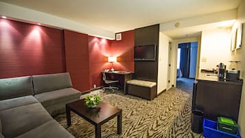 A hotel room with a sofa, a small table with a plant, a desk with a chair, a TV, and a kitchenette area.